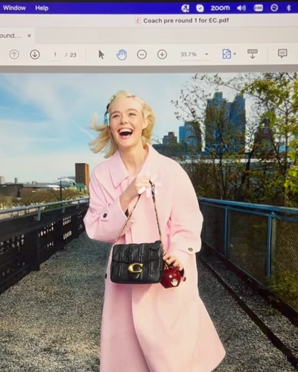 A smiling woman with blond hair wearing a pink coat and holding a black Coach purse and red apple, posing in an outdoor urban setting.