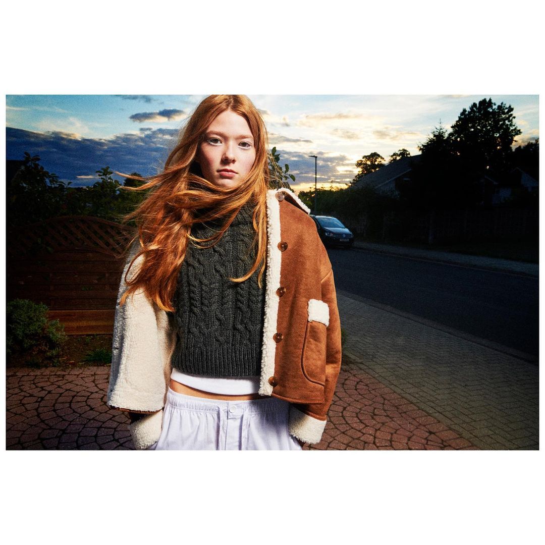 Redhead woman poses outdoors wearing a shearling-trimmed jacket, sweater, and pants on a brick walkway with an evening sky.