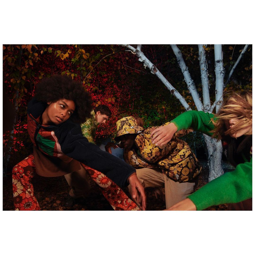 Four fashion models strike poses in an outdoor setting with foliage and white birch trees. The models are wearing patterned clothing.