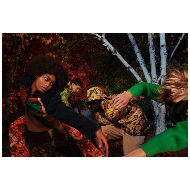 Four fashion models strike poses in an outdoor setting with foliage and white birch trees. The models are wearing patterned clothing.
