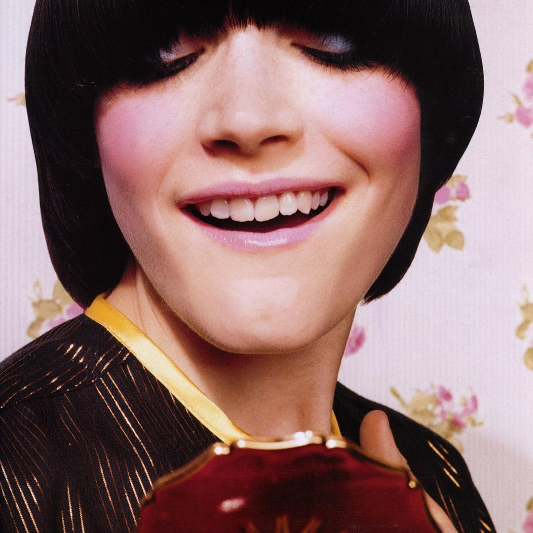 Smiling woman with blunt bangs and bright pink blush holds a maroon compact in front of a floral wallpaper backdrop in a close-up fashion shot.