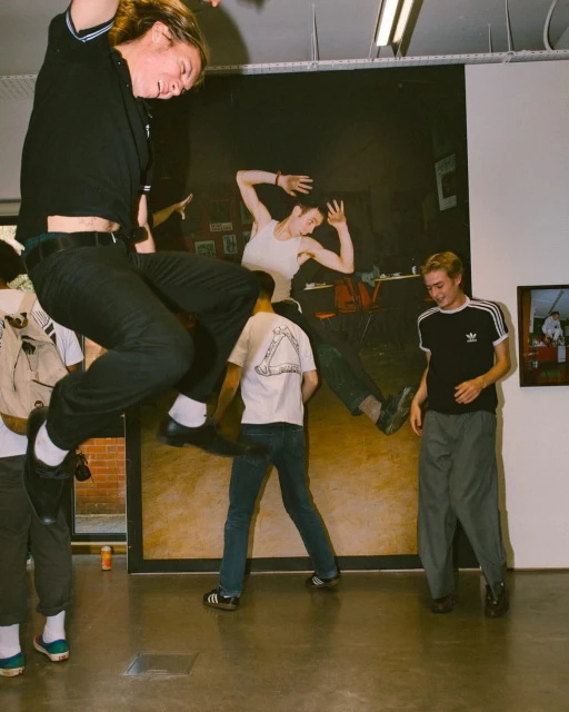 A group of young adults at an indoor art gallery, with one man jumping in front of a large artwork of a woman. 