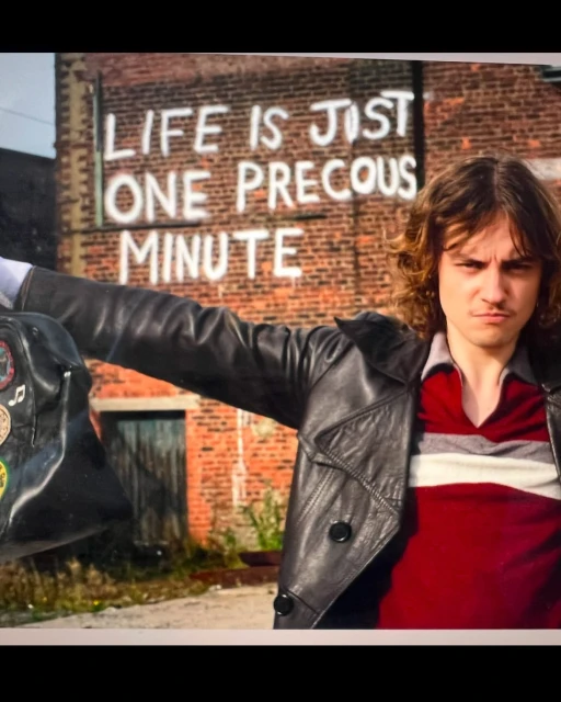 An adult male in a black leather jacket stands outdoors with a brick building behind him, featuring graffiti text and carrying a bag.