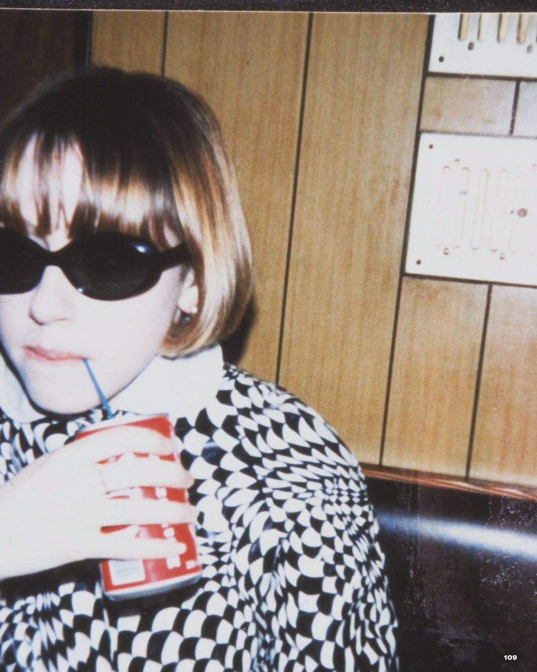 Young girl with sunglasses drinks from a can with a straw, wearing a patterned shirt, sitting in a booth with wood paneling.