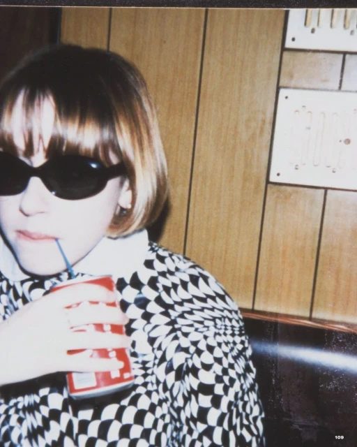 Young girl with sunglasses drinks from a can with a straw, wearing a patterned shirt, sitting in a booth with wood paneling.