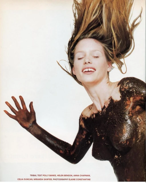 Woman covered in mud-like substance posing with open mouth and outstretched arm against a white backdrop with long, blonde hair.