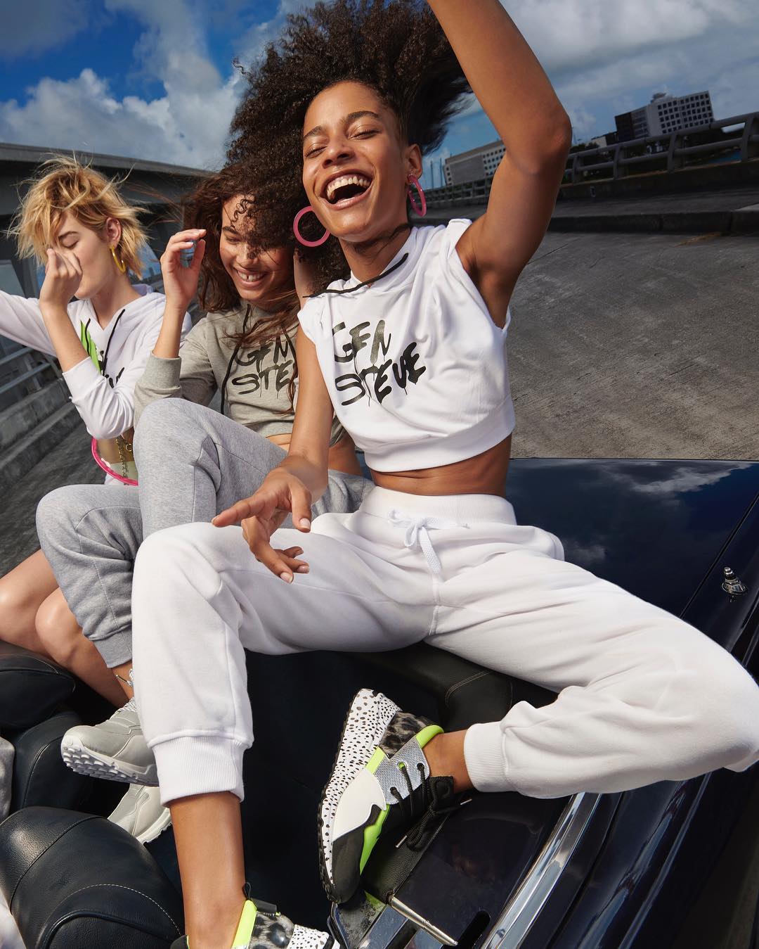 Three young adult women pose in an open convertible in casual clothes, smiling and laughing against an overcast sky in an outdoor setting.