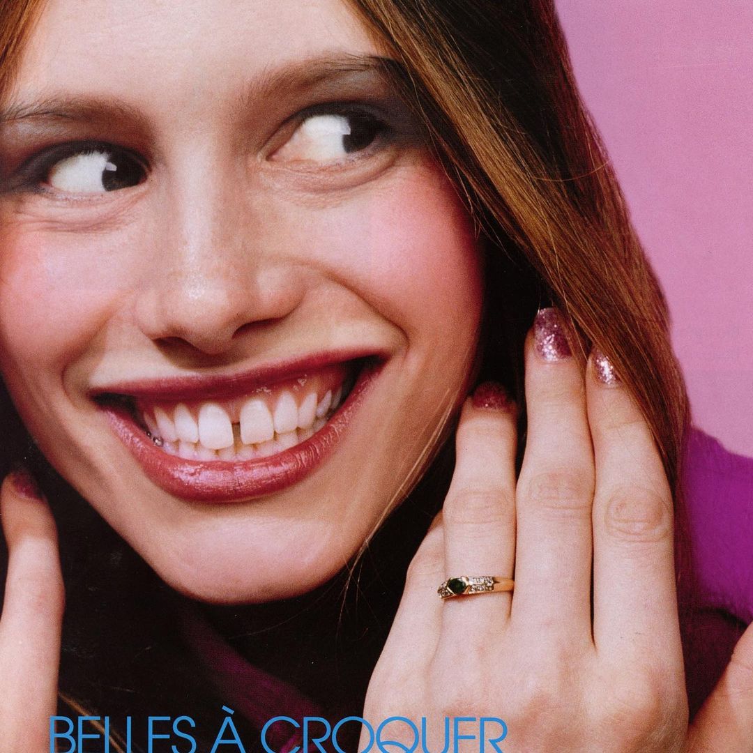 Close-up of a smiling young woman with auburn hair, pink glitter nails, and a ring on her finger, against a pink background.