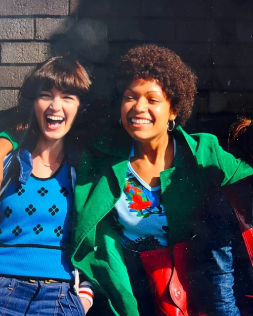 Two women smiling outdoors, wearing colorful tops and jackets, with one woman having an afro hairstyle, standing against a brick wall.