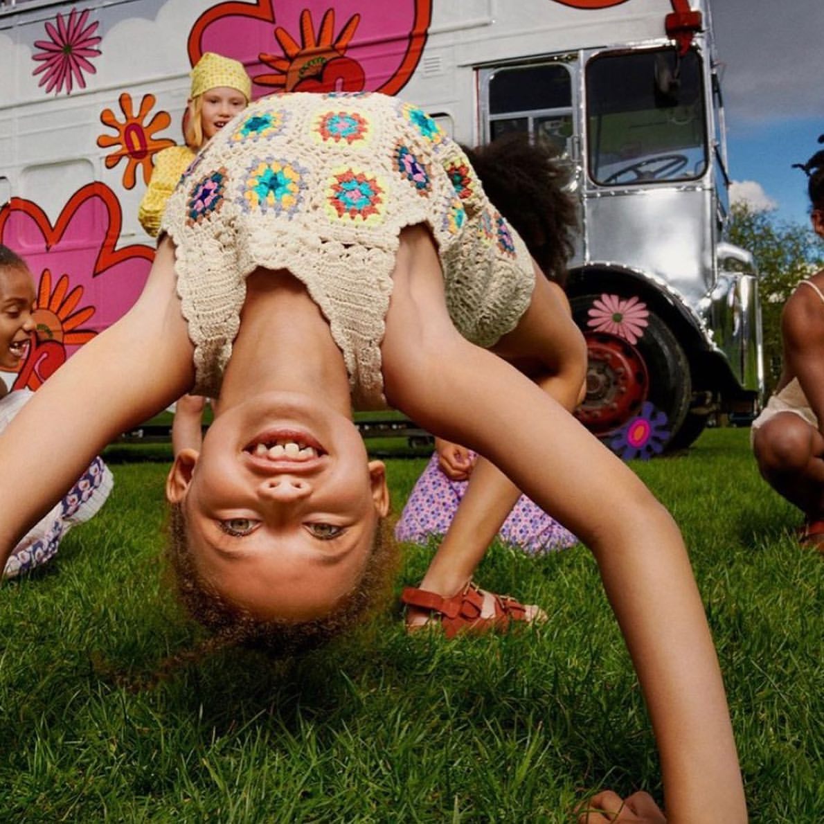 Child in a crocheted dress doing a backbend on grass with other children and a van with floral decoration in the background.