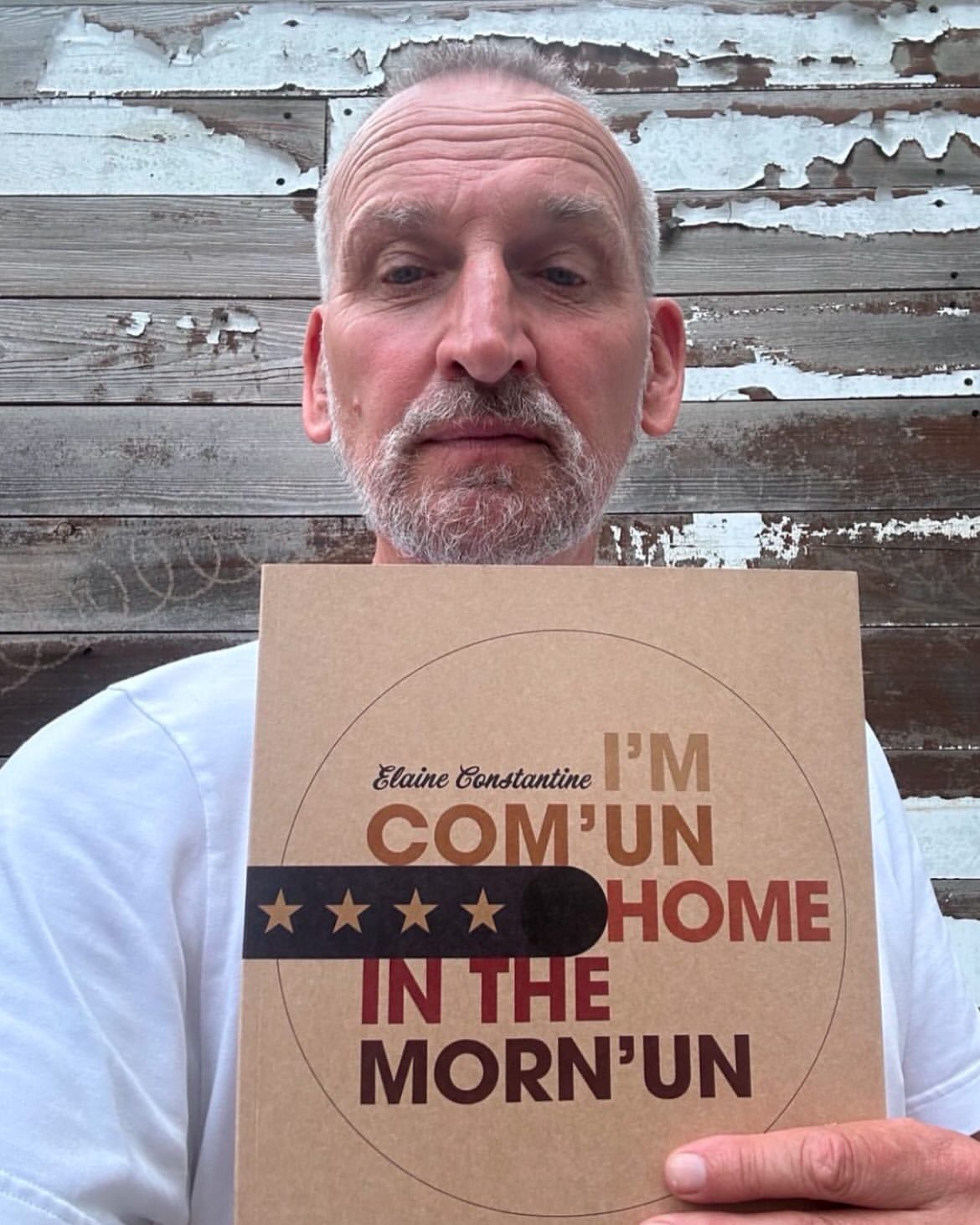 Close-up of mature man holding a book titled 'Com'un Home in the Morn'un' against a rustic weathered wood background.