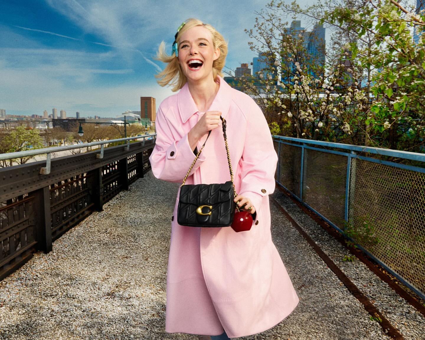 Elle Fanning wears a pink coat, carrying a black quilted purse with a gold chain, standing on a pedestrian bridge in an urban park.
