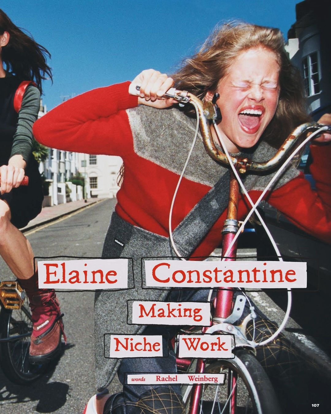 Two women riding bicycles on a city street, one with a very expressive face, wearing a red sweater with gray accents.