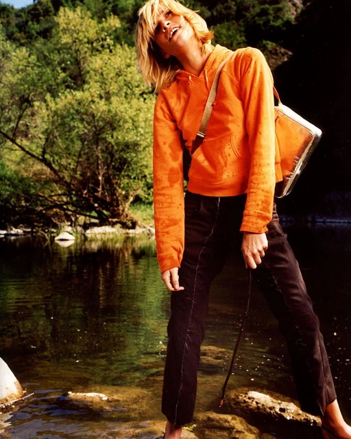 Adult female standing in a river with a fishing rod, wearing an orange hoodie and dark jeans, with a green forested background.