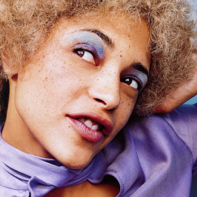 Portrait of a woman with curly blonde hair and blue eyeshadow wearing a periwinkle collared shirt looking up and smiling slightly.