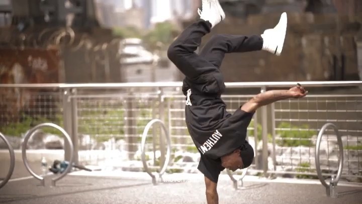 Athletic man performs handstand on public walkway with urban background, wearing black athletic wear and white sneakers, showcasing fitness and strength.