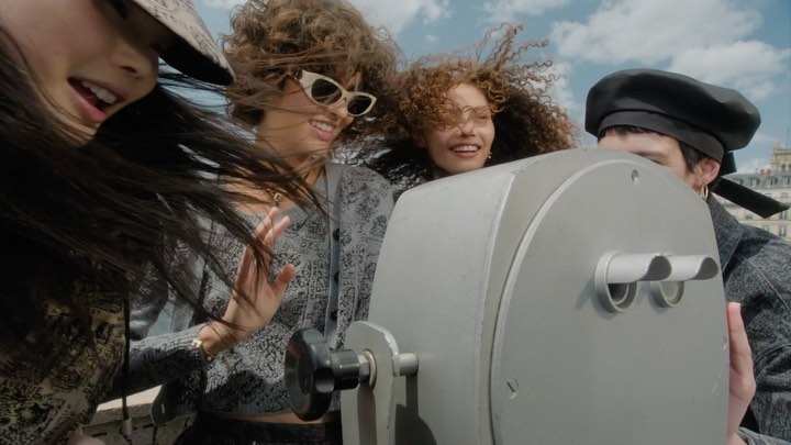 Four smiling people, including three women and one man, look at a coin-operated telescope with city buildings and a cloudy sky in the distance.