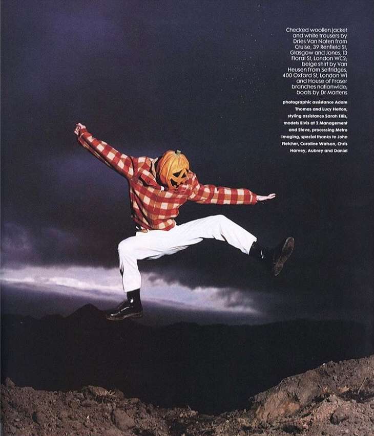 Person in a pumpkin head costume jumping in the air at night near mountains, wearing a checkered jacket and white trousers.