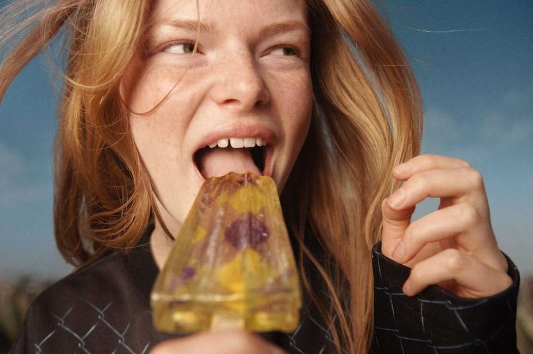 Close-up of a young woman with red hair eating a clear popsicle with flowers, set against a blue sky backdrop.