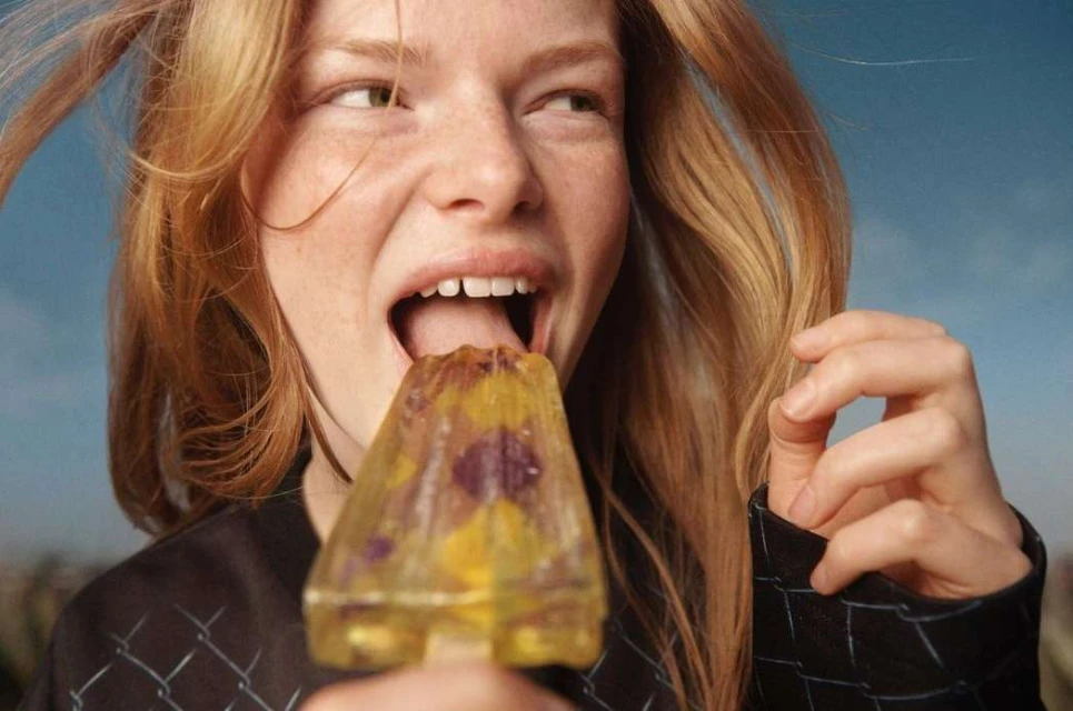 Close-up of a young woman with red hair eating a clear popsicle with flowers, set against a blue sky backdrop.