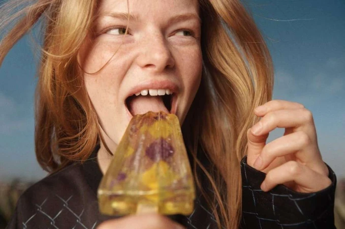 Close-up of a young woman with red hair eating a clear popsicle with flowers, set against a blue sky backdrop.