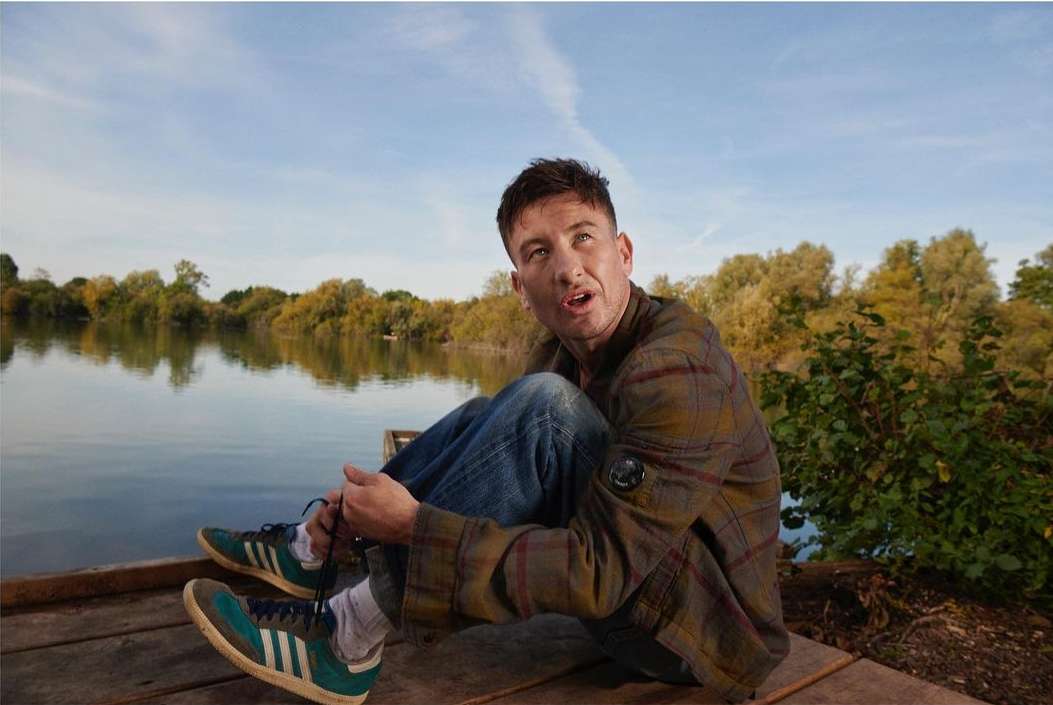Adult male sitting on a dock near a lake wearing jeans, a plaid shirt, and sneakers in an outdoor setting on a sunny day.