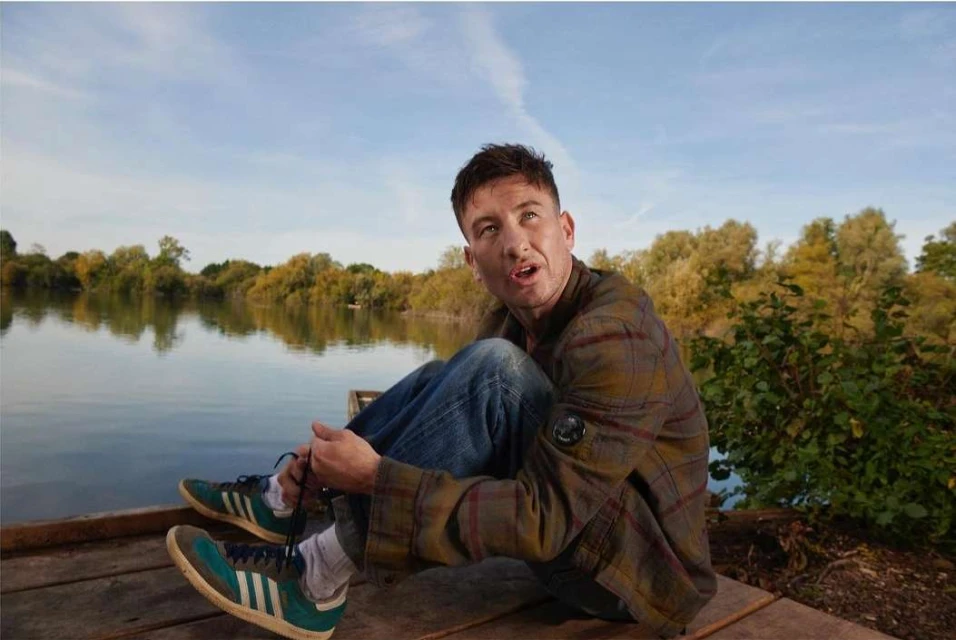 Adult male sitting on a dock near a lake wearing jeans, a plaid shirt, and sneakers in an outdoor setting on a sunny day.
