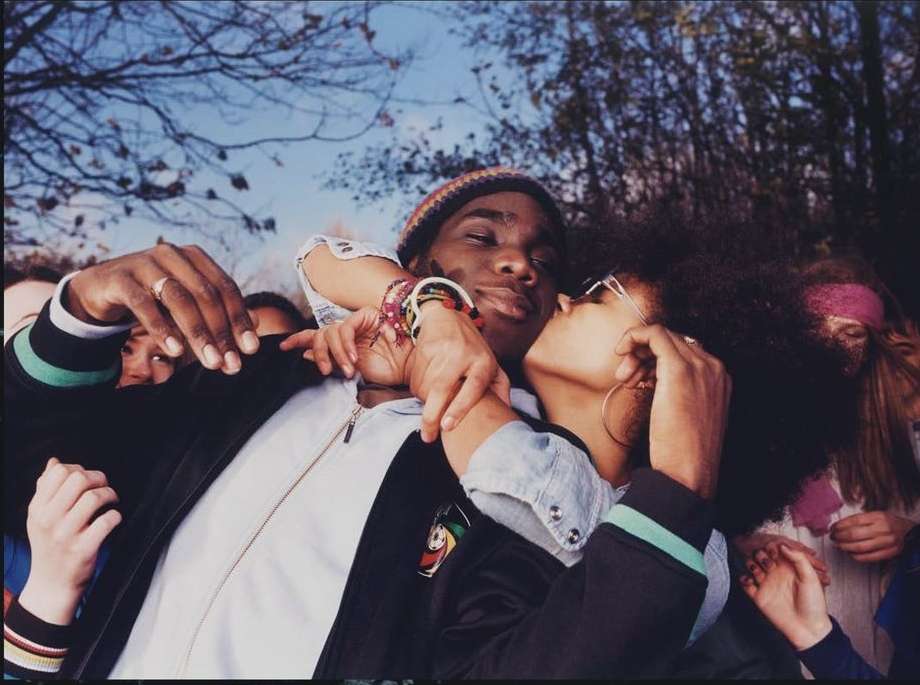 Young woman kissing a young man on the cheek amid a crowd outdoors under bare trees, captured by Elaine Constantine.