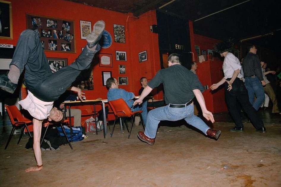 Indoor scene shows a man doing a handstand in front of a crowd of people gathered inside with red walls and overhead lighting.