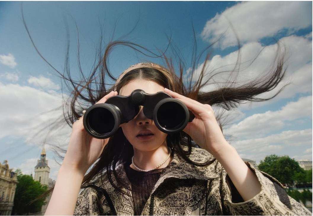 A young woman in a patterned jacket looks through binoculars, standing outside with a blue sky and buildings in the background.