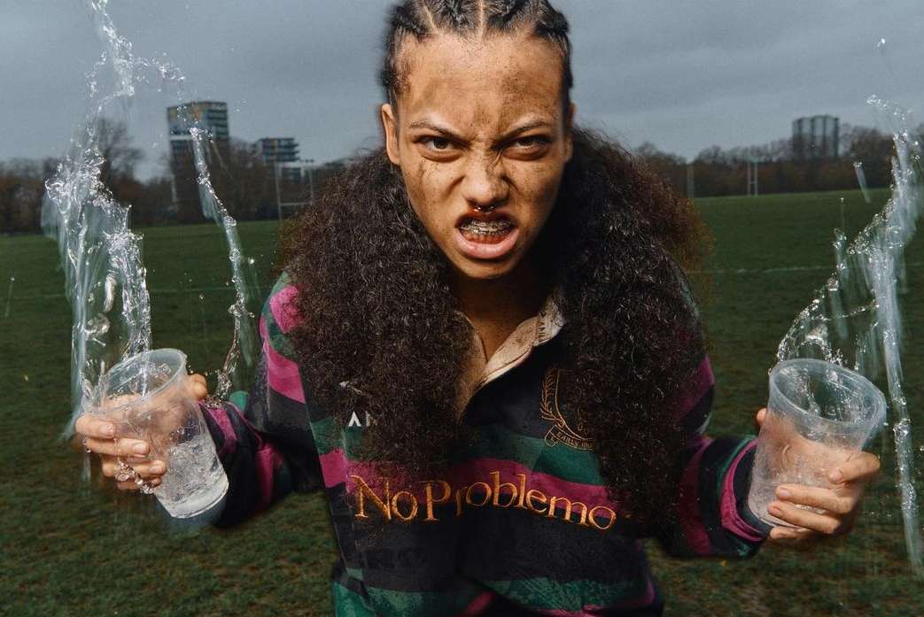 Athletic woman with braces stares intently, holding glasses of water in an outdoor field setting under a cloudy sky, wearing a rugby shirt.