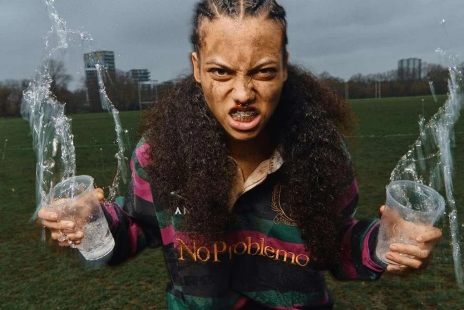 Athletic woman with braces stares intently, holding glasses of water in an outdoor field setting under a cloudy sky, wearing a rugby shirt.