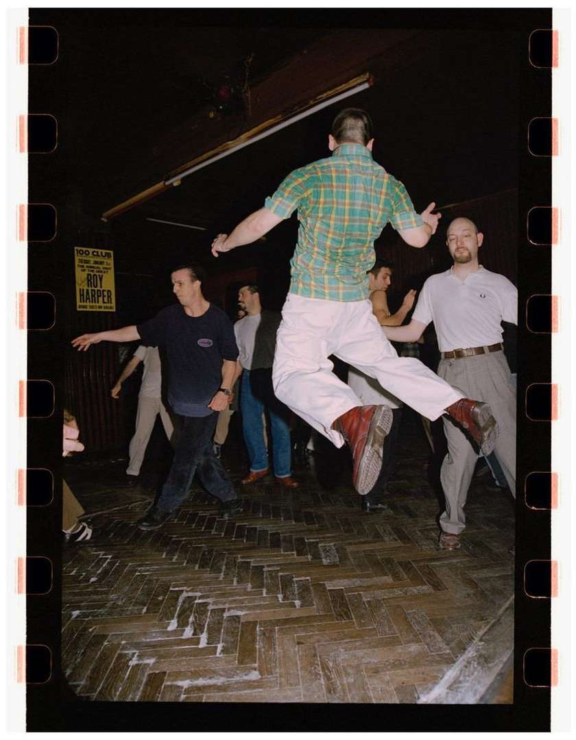 Man jumping in a crowded dance hall, captured on film with visible film border; dancers and parquet floor in the background; yellow poster visible.
