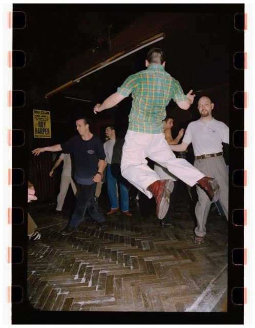 Man jumping in a crowded dance hall, captured on film with visible film border; dancers and parquet floor in the background; yellow poster visible.