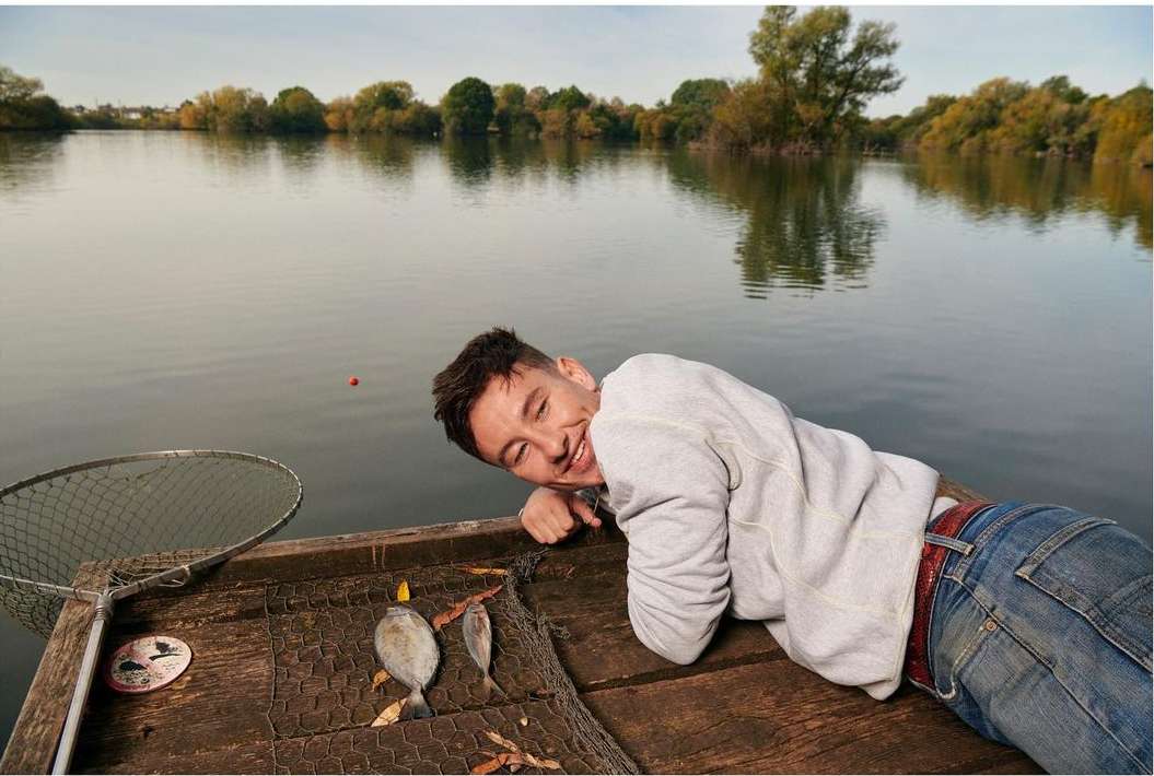 Smiling adult male lying on a wooden pier next to caught fish, fishing net, with a lake and trees in the background.