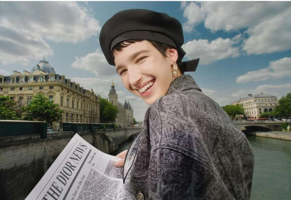 Smiling woman with dark beret in Paris holding 'The Dior News' newspaper in front of city buildings and bright blue cloudy sky.