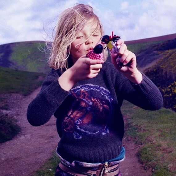 A blonde child in a patterned sweater stands outdoors holding up two small yarn dolls, with a hillside in the blurred background.