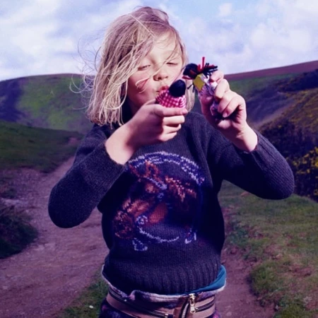 A blonde child in a patterned sweater stands outdoors holding up two small yarn dolls, with a hillside in the blurred background.