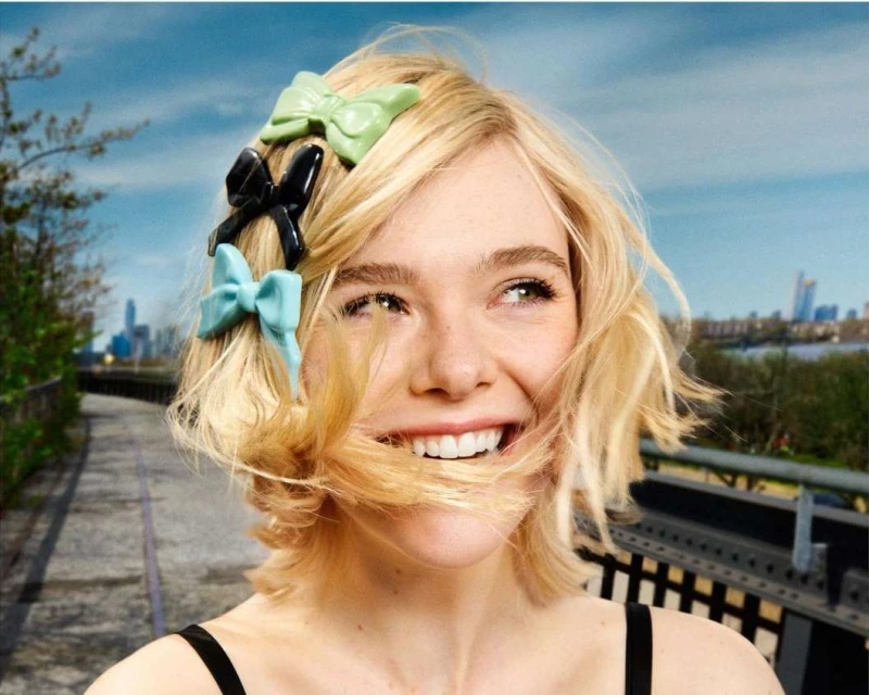 Smiling woman with blonde hair adorned with multiple hair bows of varying colors, posing outdoors with a city skyline in the distance.