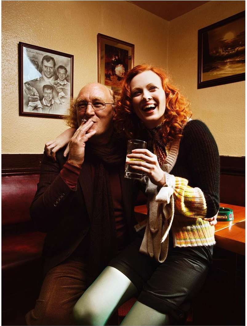 Couple in pub: Mature man with cigarette, woman with red hair laughing while holding drink, in warm lighting, indoor.
