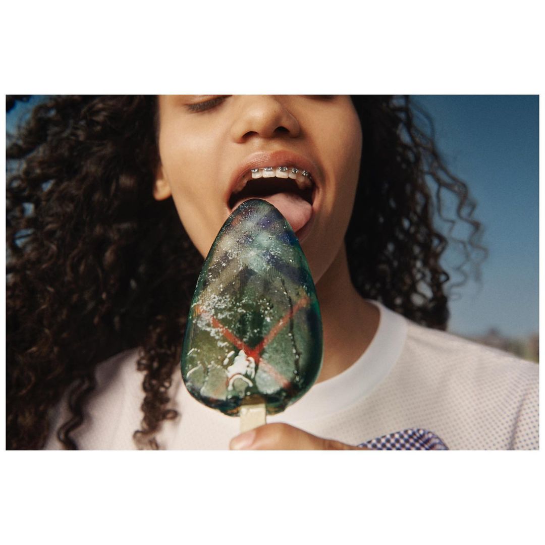 A person with curly hair and braces puts their tongue to a green and blue ice pop against a bright blue sky in a close-up shot.