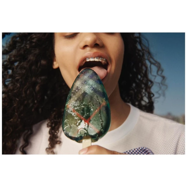 A person with curly hair and braces puts their tongue to a green and blue ice pop against a bright blue sky in a close-up shot.