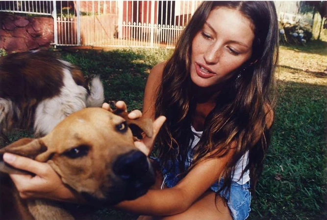 Adult woman with long hair petting a brown dog in a backyard setting; lush green grass, white fence, and a red-rocked wall.