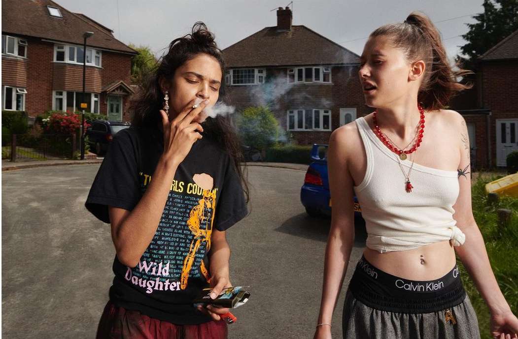 Two young women outdoors in casual clothing, one smoking a cigarette, with suburban houses and a blue car in the background.