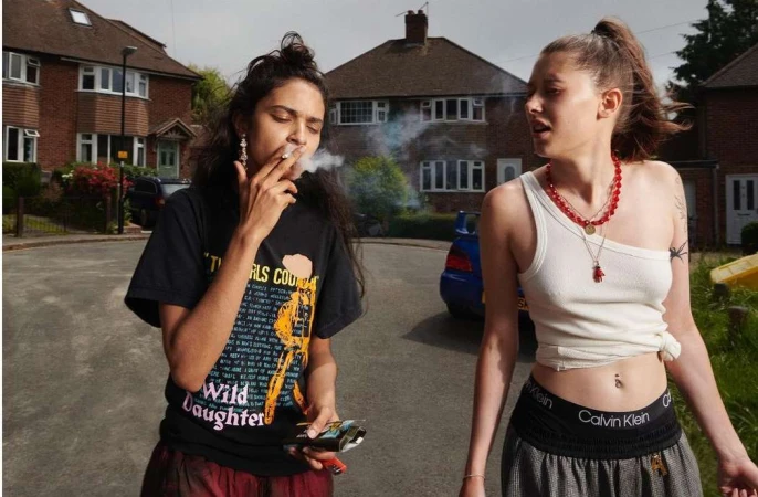 Two young women outdoors in casual clothing, one smoking a cigarette, with suburban houses and a blue car in the background.