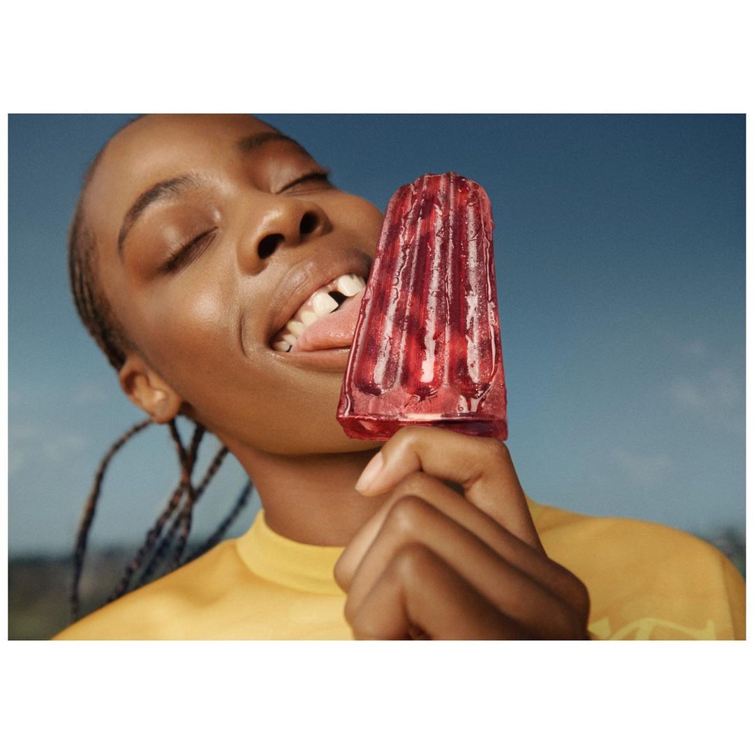 Young Black woman eating a red ice pop outdoors against a blue sky background, wearing a yellow t-shirt with braided hair.