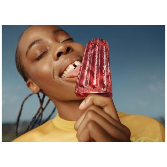 Young Black woman eating a red ice pop outdoors against a blue sky background, wearing a yellow t-shirt with braided hair.