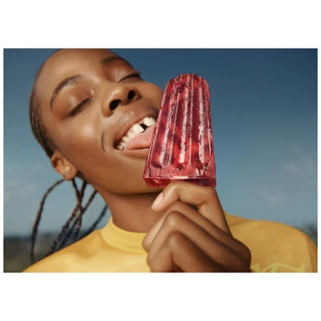 Young Black woman eating a red ice pop outdoors against a blue sky background, wearing a yellow t-shirt with braided hair.