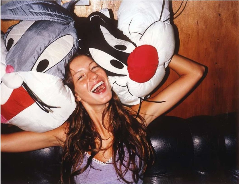 Smiling adult woman posing with Bugs Bunny and Sylvester the Cat plush toys against a wall and leather couch in an indoor setting.