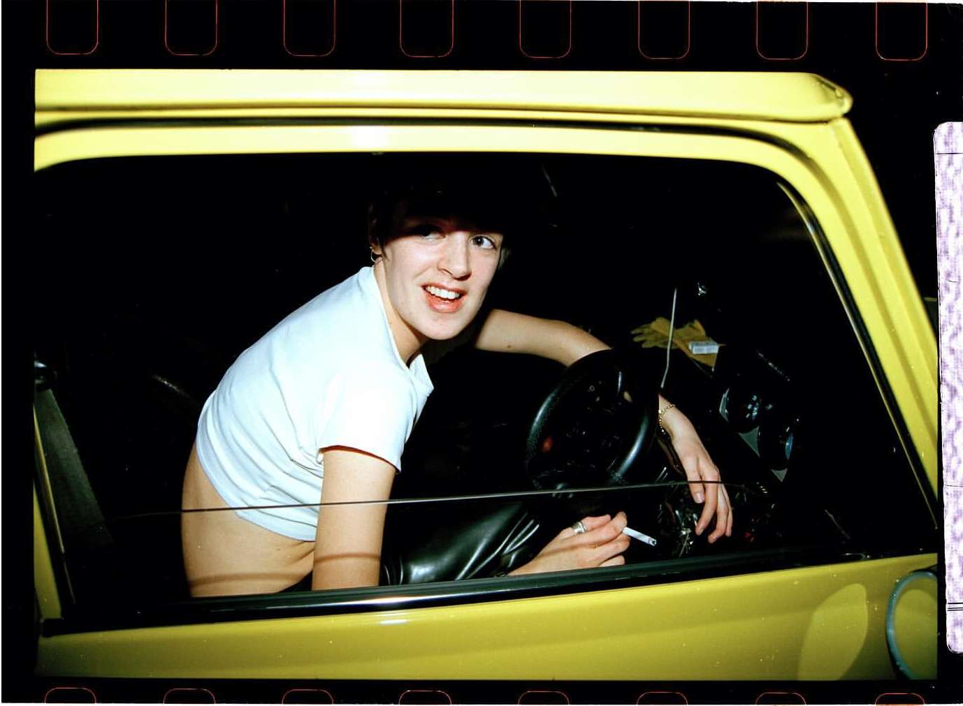 Woman in a yellow car at night, looking toward the camera with a cigarette in hand and wearing a white t-shirt.